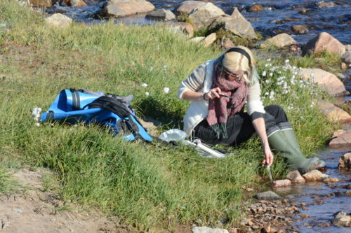 Kat sits in a grassy field close to a shallow river, measuring sounds and making field recordings.