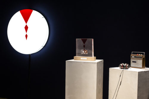 Against a dark backdrop, a white circular fixture on the left side of the wall is lit up, with an inverted red triangle at the top of it and two smaller red diamonds below it, moving towards the center of the circle. In the middle of the photo is a cassette tape suspended in a clear plastic mold that has the same triangle and diamond pattern, which the words "Good Luck" printed on the wooden block holding it up. At the very right is an old radio with a set of wired headphones connected to it.